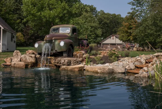 An old, rusted pickup truck is positioned on rocks, with water flowing from its front into a pond, expertly arranged by Valparaiso landscapers in a landscaped yard with trees and houses in the background.