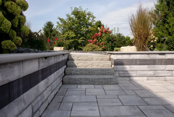 Stone steps ascend from a paved patio, bordered by low retaining walls, to a lush garden of flowering plants and shrubs—expertly crafted by Crown Point landscaping professionals.