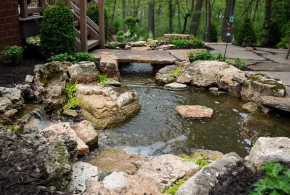A small backyard pond with rocks and green plants, designed by Valparaiso landscapers, featuring a stone bridge and surrounded by a patio and trees in the background.